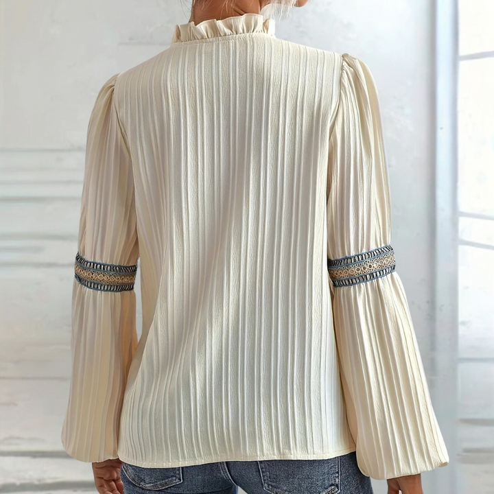 Crèmekleurige blouse met lange geplooide mouwen en blauwe borduursels, gedragen door een persoon, tegen een lichte achtergrond.