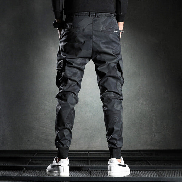 Zwarte cargobroek voor heren, met meerdere zakken en elastische boorden, gedragen met witte sneakers. Stijlvolle streetwear mode.